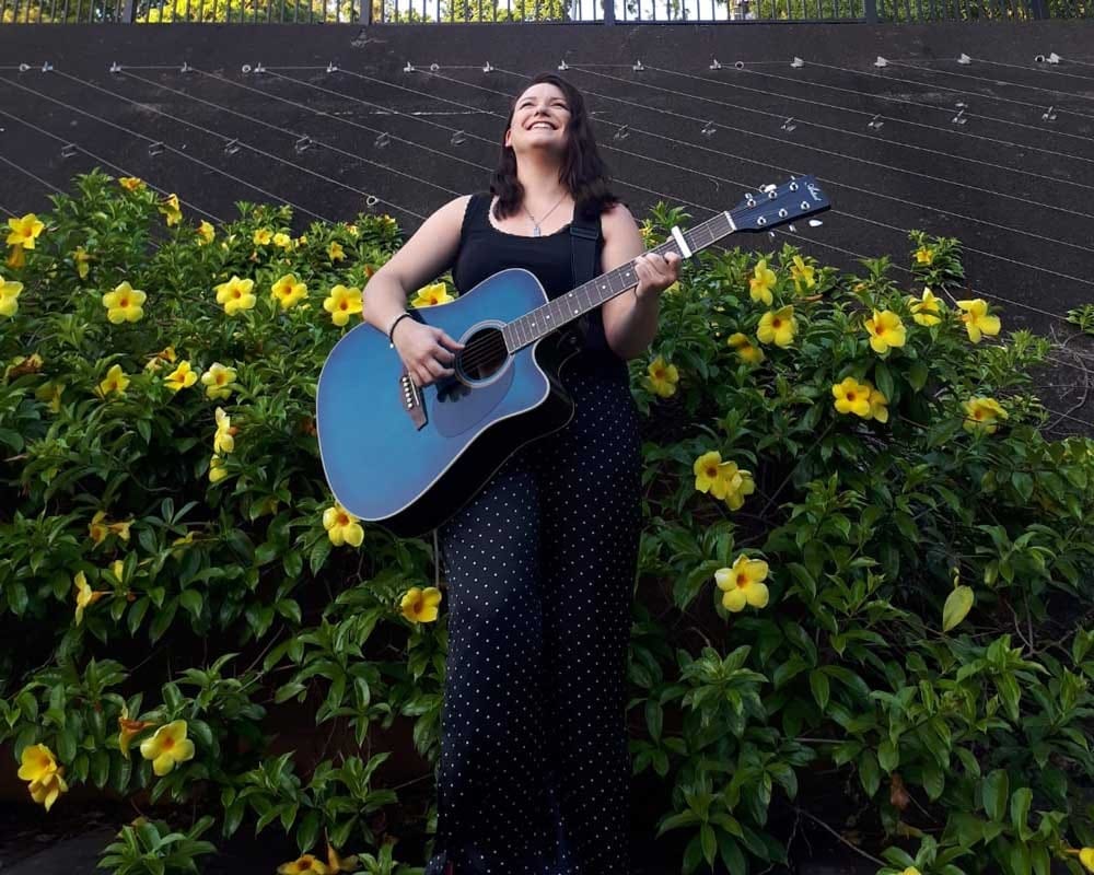 Trabajar en Australia chica morena con un vestido largo negro y una guitarra de color azul y detrás flores de color amarillo